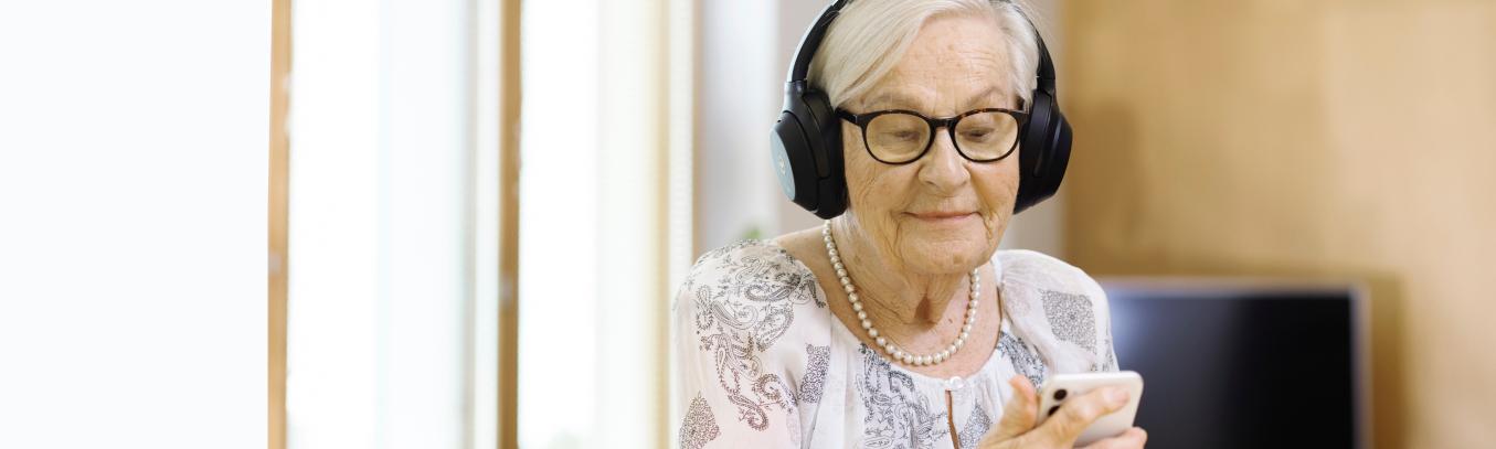 Seniori-ikäinen nainen kännykkä kädessä ja luurit päässä.